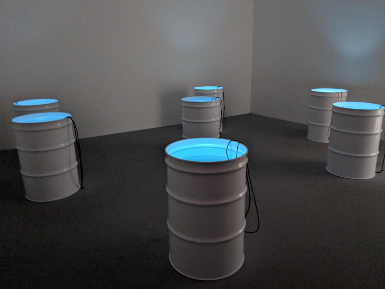 seven white oil drums with blue light and two black wires coming out of each