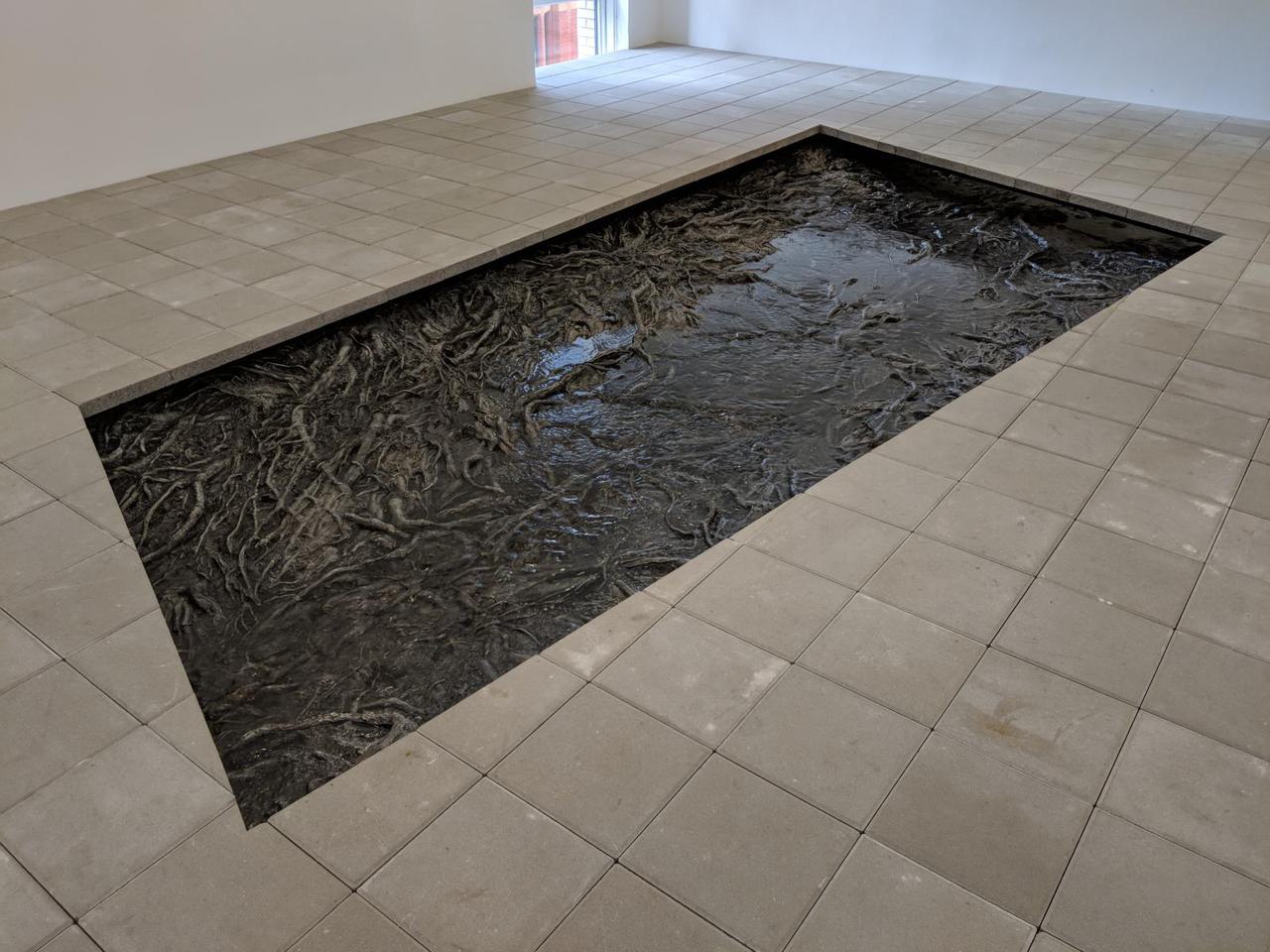 stone tile floor with large rectangular hole in the center, with a metal surface that looks organic, like tree branches or roots.