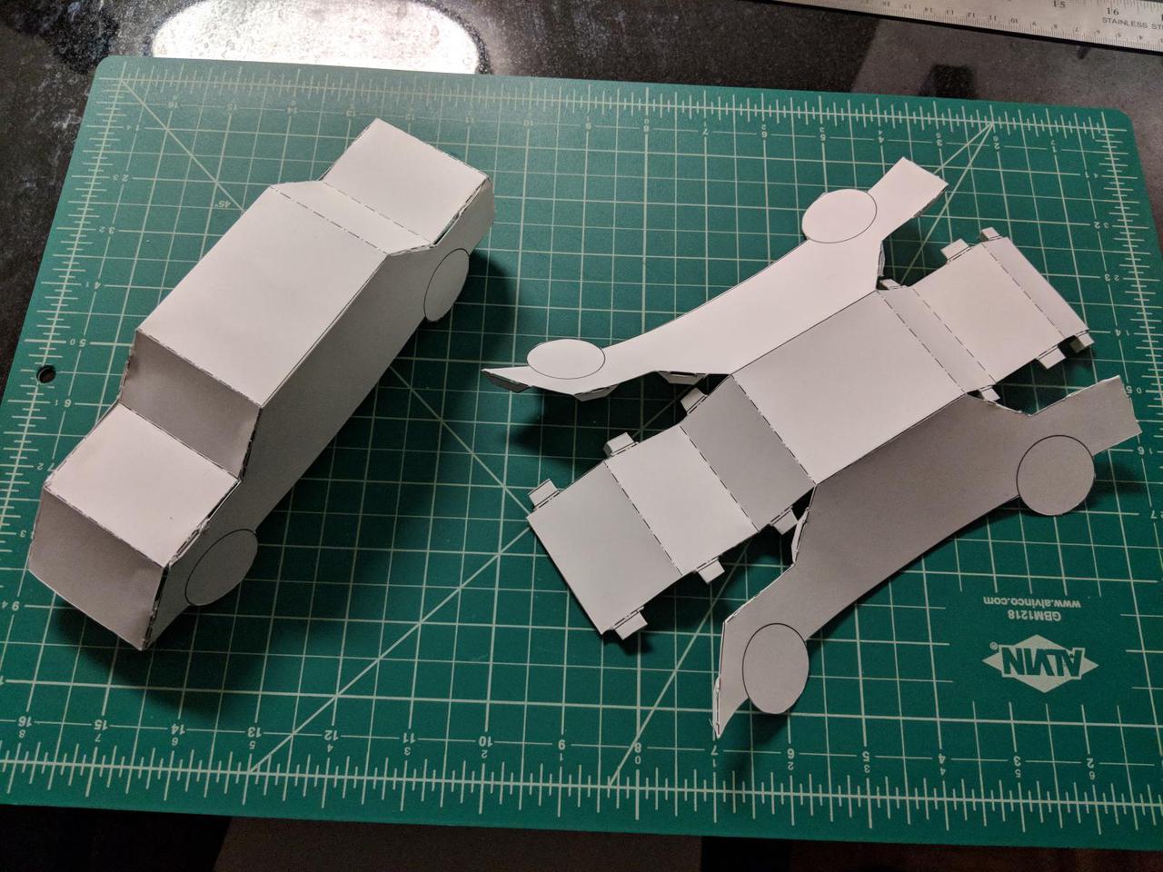 Two paper cars on a green cutting mat. One is assembled and the other is waiting to be assembled. Both are made of white paper.