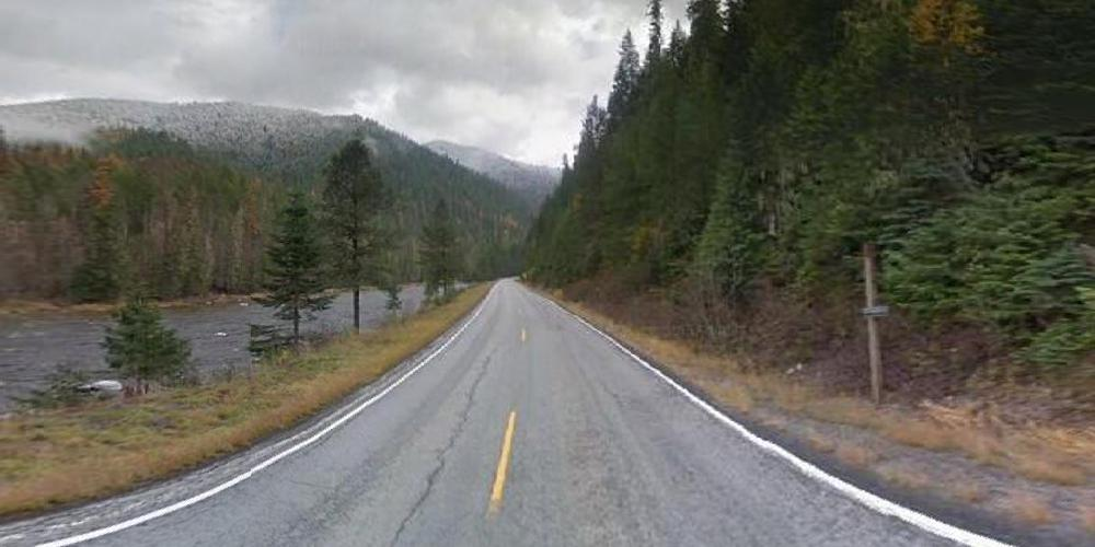 2 lane highway with stream to the left and trees to the right and clouds in the sky.