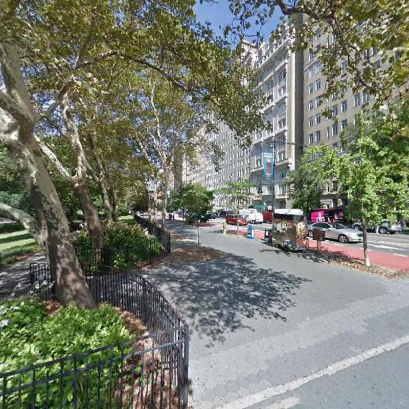 NYC scene with trees the foreground, a sidewalk, and a road and a building in the background. The sky is blue.