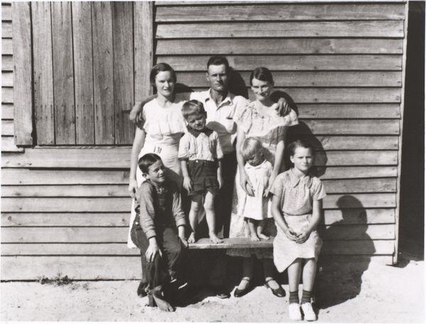 old photograph of family standing in front of old wooden house.