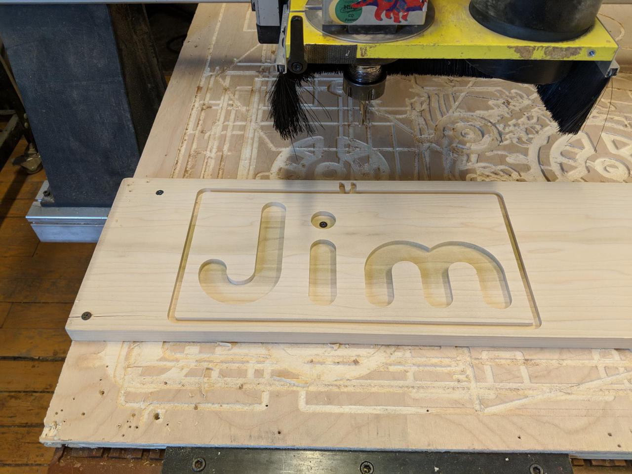 wood attached to CNC bed with the letters J I M carved into it. The CNC bit is hovering above it.