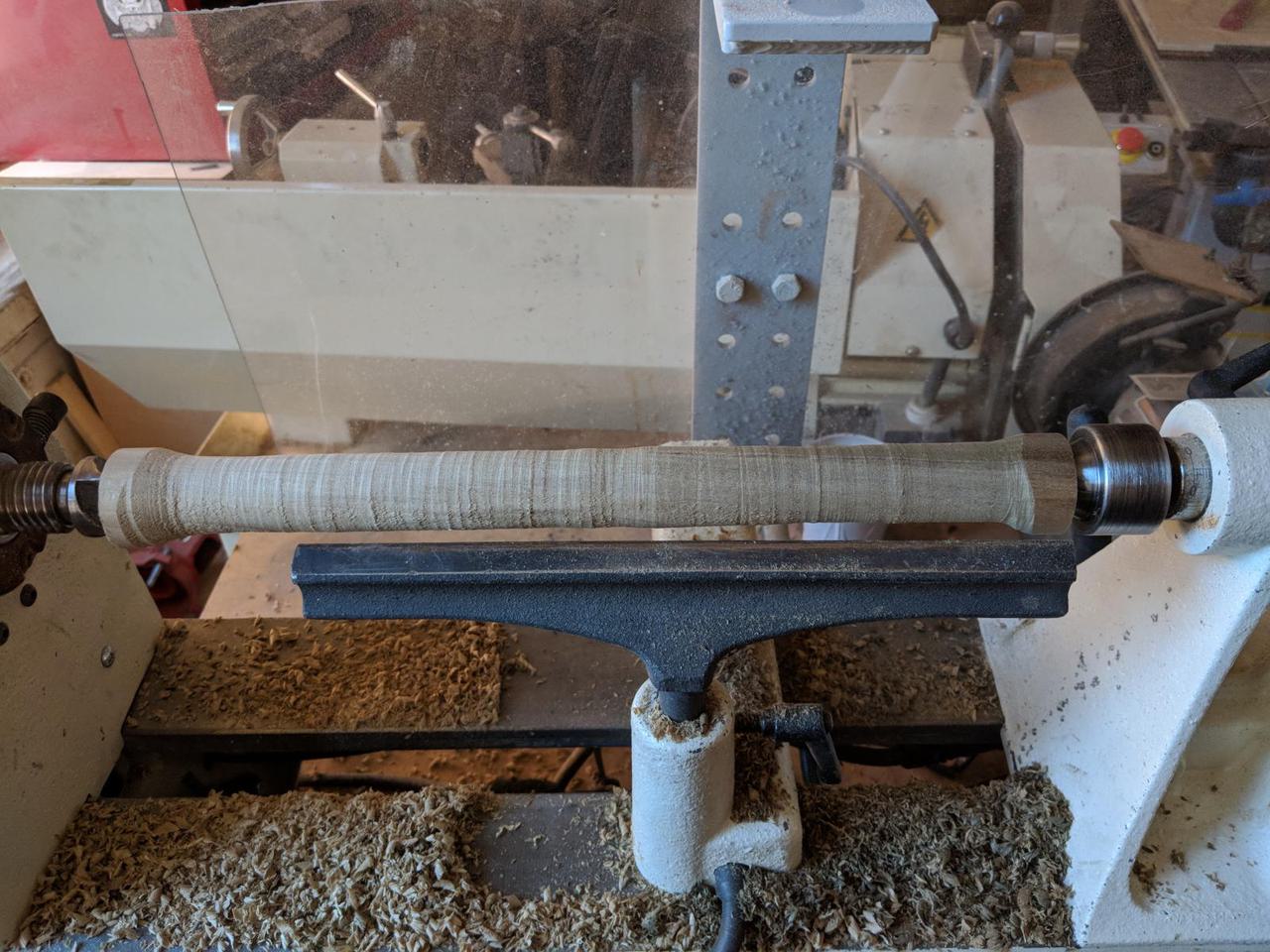 wooden cylinder mounted in wood lathe, with cylinder thinner than before and sawdust everywhere.