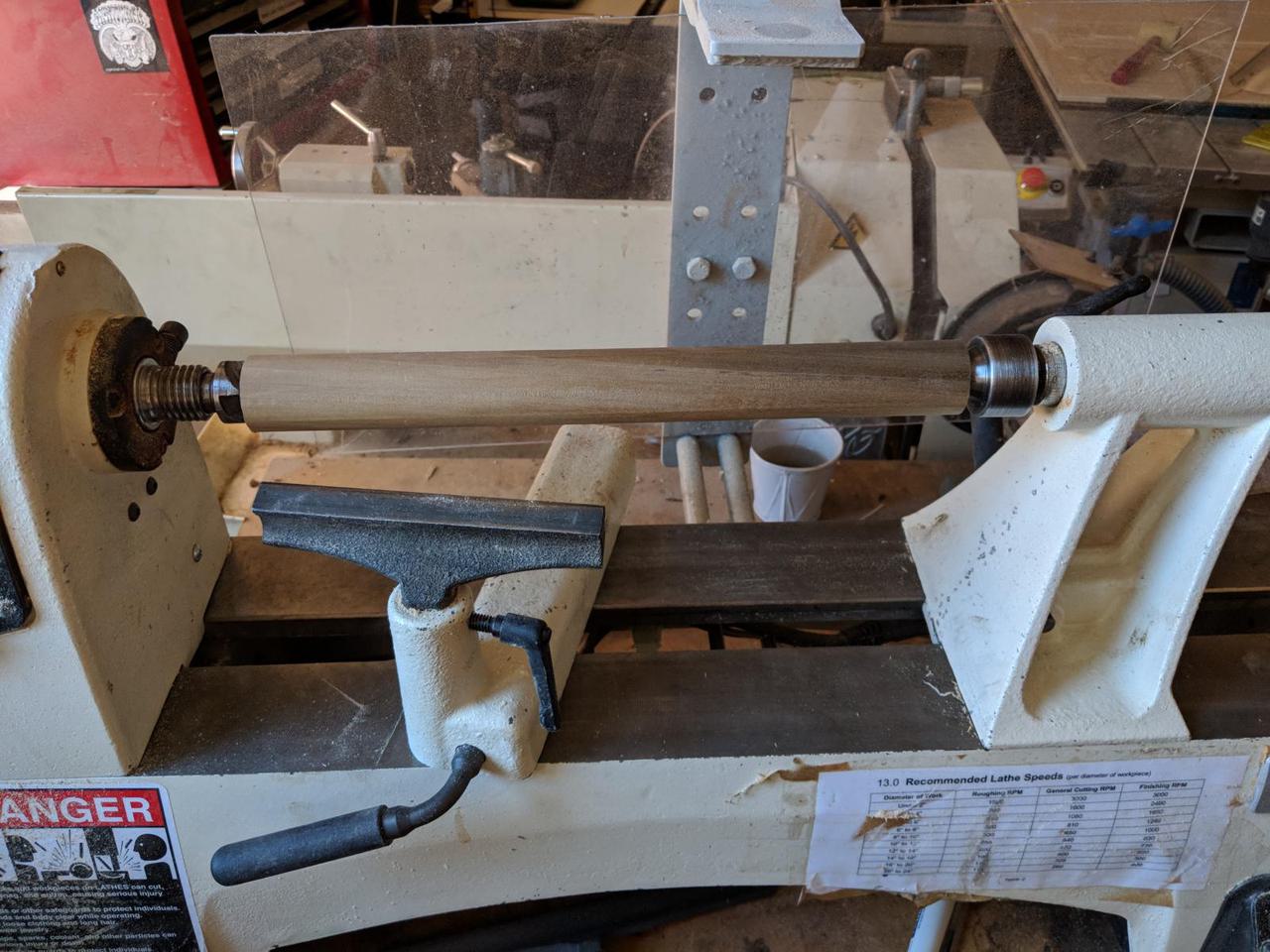 wooden cylinder mounted in wood lathe, held in place with both ends hooked into the machine.