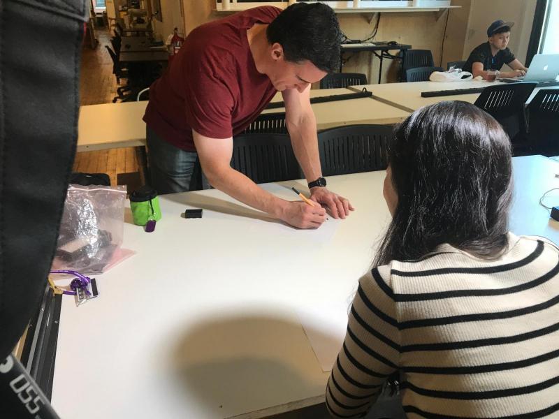 Me leaning over a table drawing with a pen on a piece of paper. Yeonhee is facing the table in the other direction.