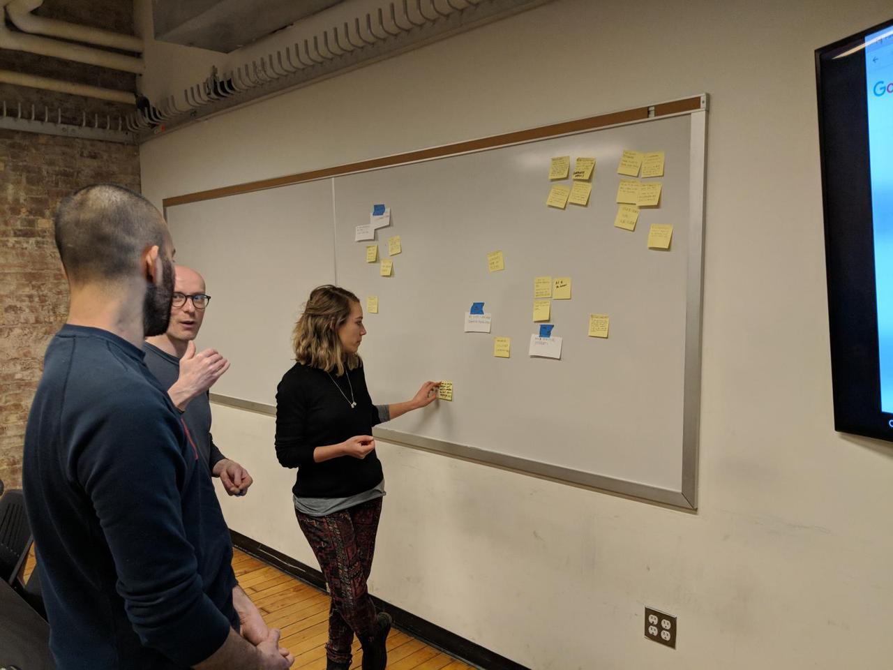 three ITP students facing a whiteboard with many postit notes attached to it