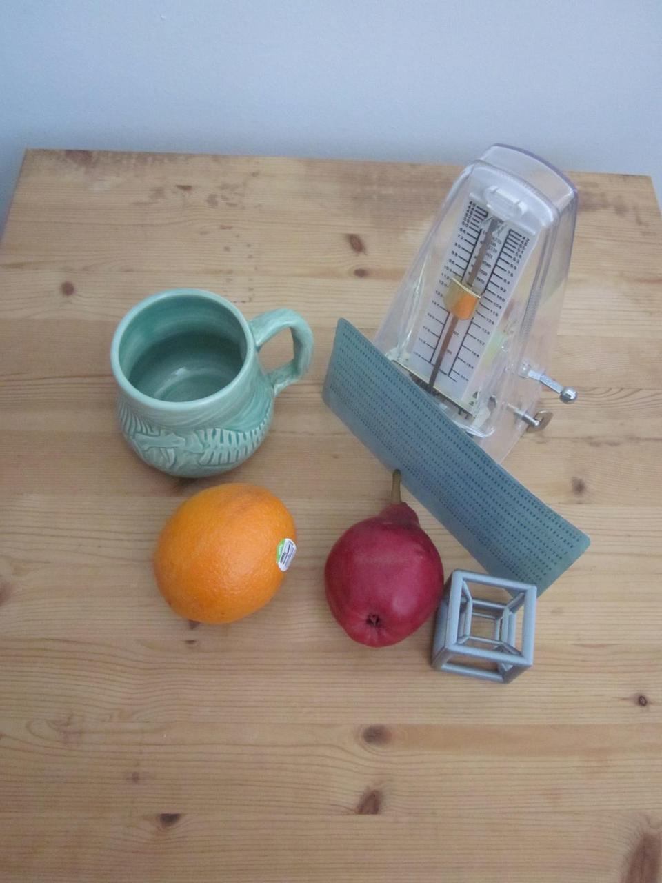 still life of an orange, an apple, a 3D printed hypercube, a green mug, a metrodome, and a blue card, all sitting on a wooden board