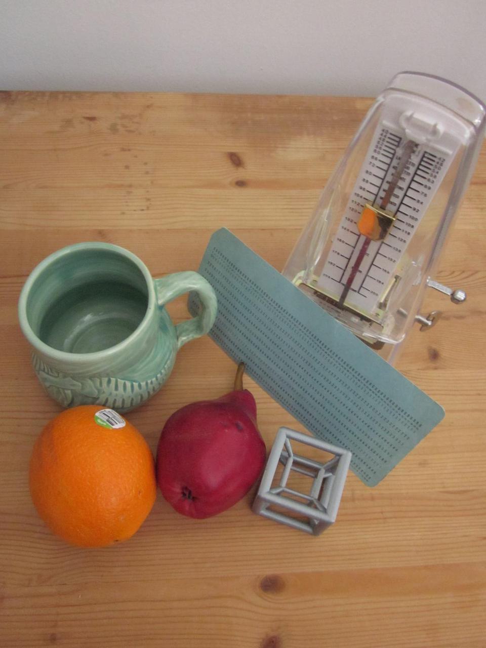 still life of an orange, an apple, a 3D printed hypercube, a green mug, a metrodome, and a blue card, all sitting on a wooden board