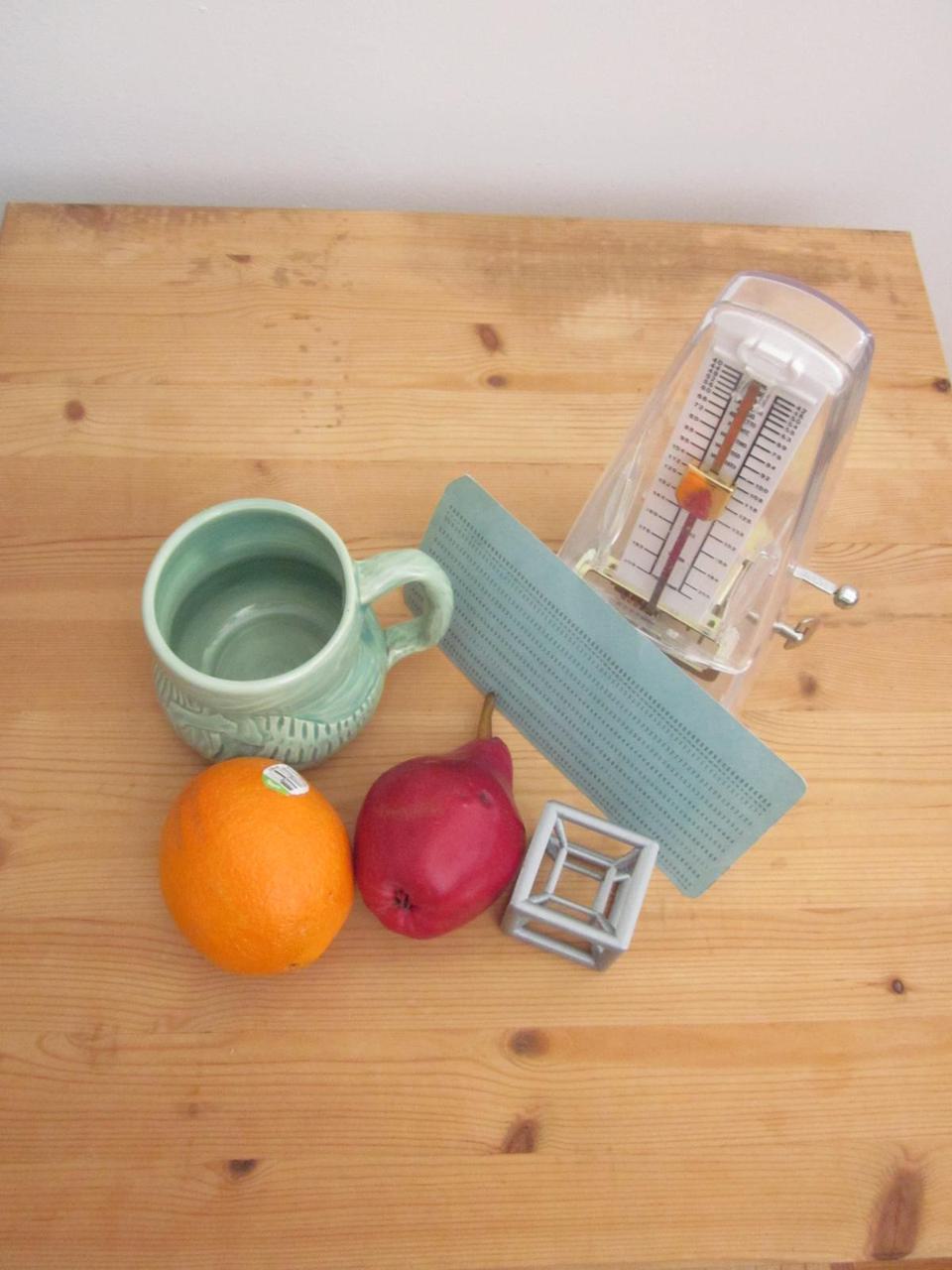 still life of an orange, an apple, a 3D printed hypercube, a green mug, a metrodome, and a blue card, all sitting on a wooden board