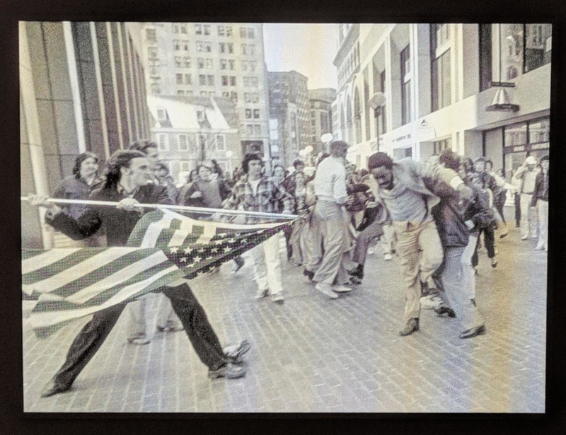 Hank Willis Thomas' work featuring retroflective technology, activated by a camera flash. One image shows a white man holding an American flag in a menacing manner, about to hit an African American man. The other image shows just the flag.