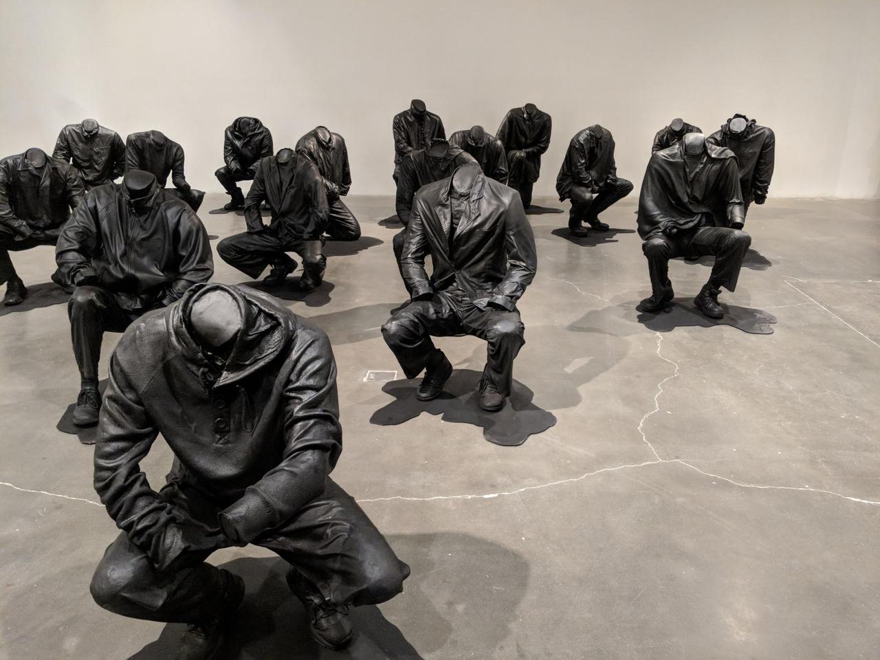 Seventeen human sculptures, each kneeling or about to kneel, all on a concrete floor.