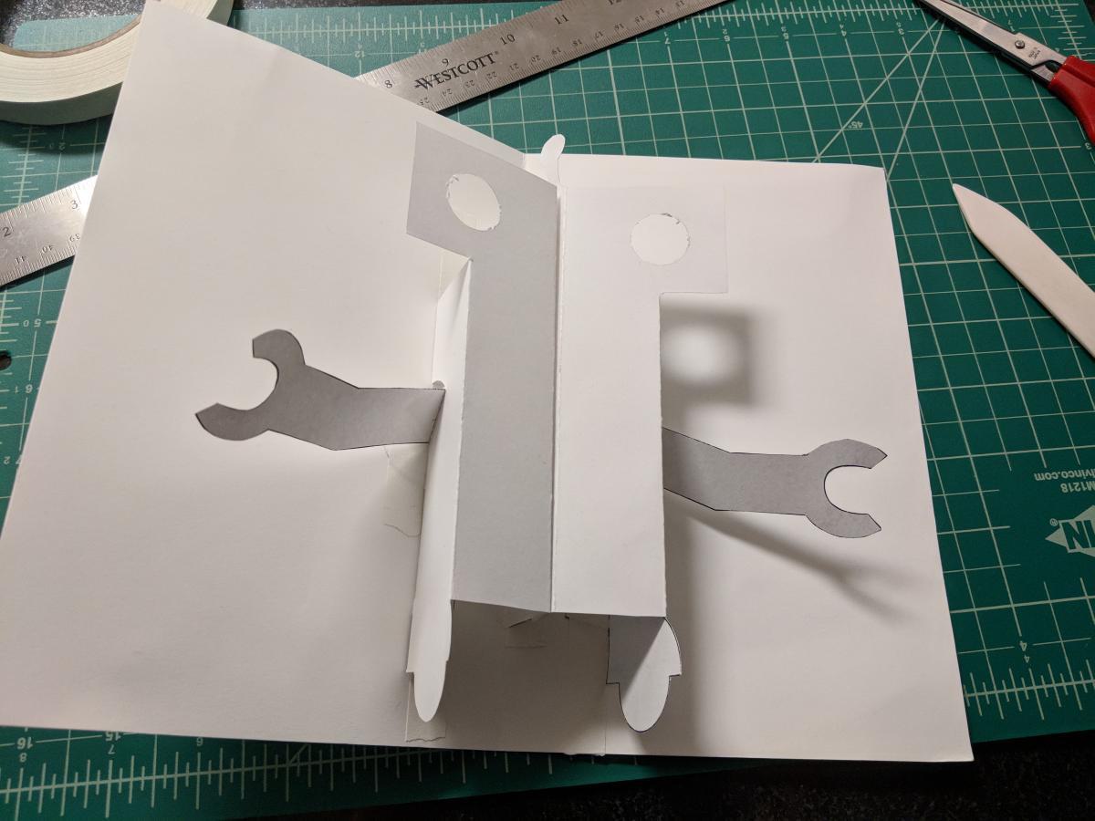 folded paper card with a raised rectangular surface in the center, some paper arms sticking out the side of the raised surface, and two holes at the top for eyes.