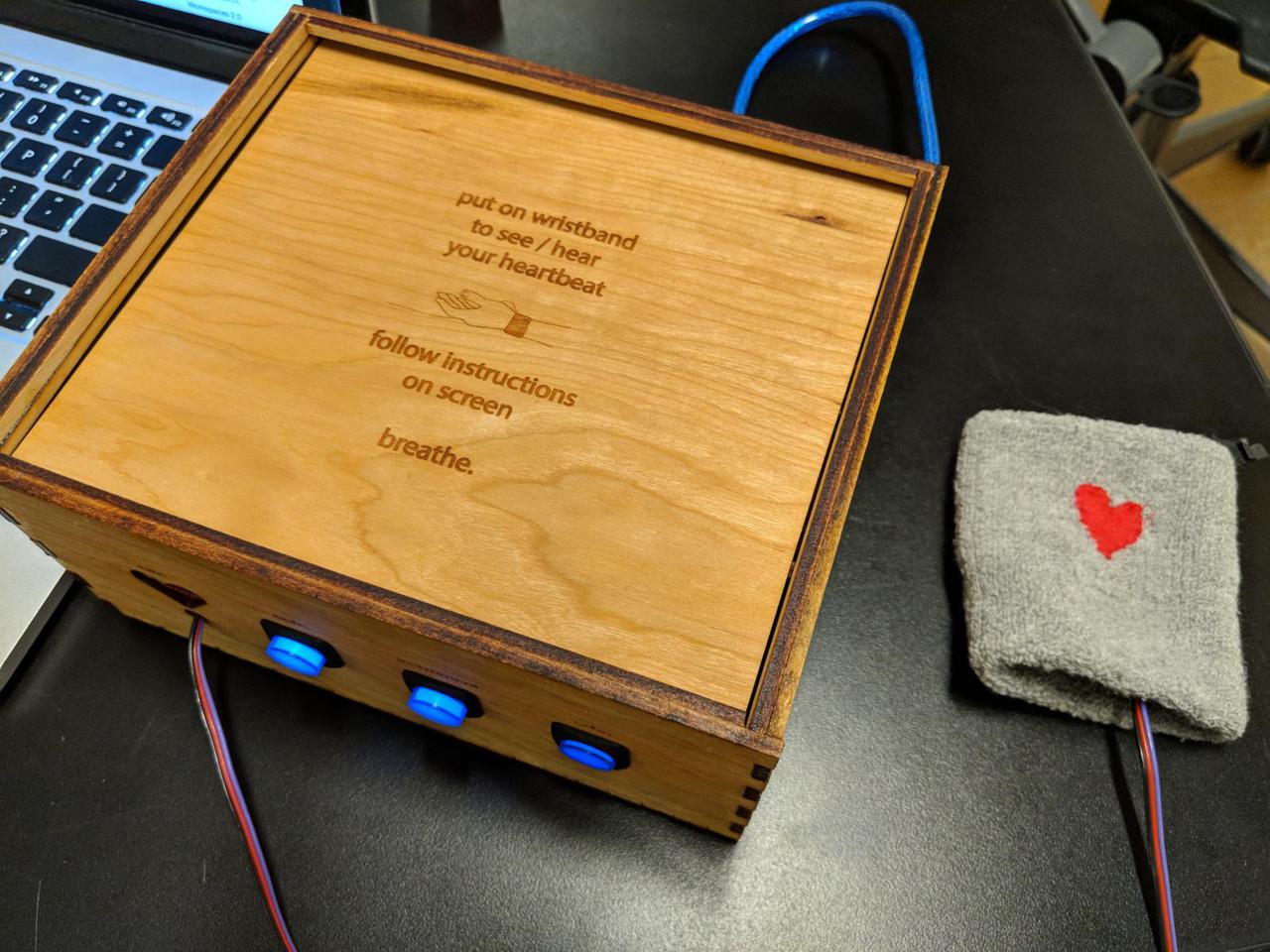 wooden box with 3 lit up buttons on front, and wire coming out of it leading to a pulse sensor attached to the inside of a wrist band.
