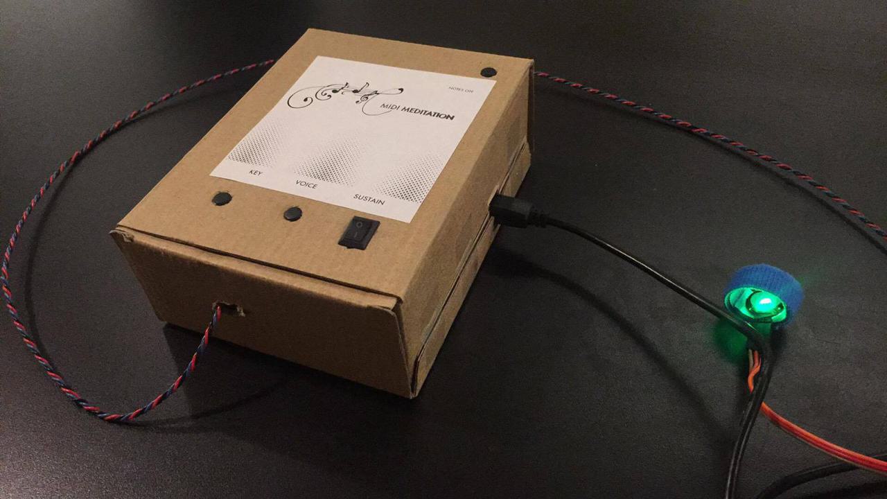 cardboard box with MIDI meditation printed on the top, and three buttons labeled key, voice, and sustain, with pulse sensor wire coming out of it and pulse sensor lit up.