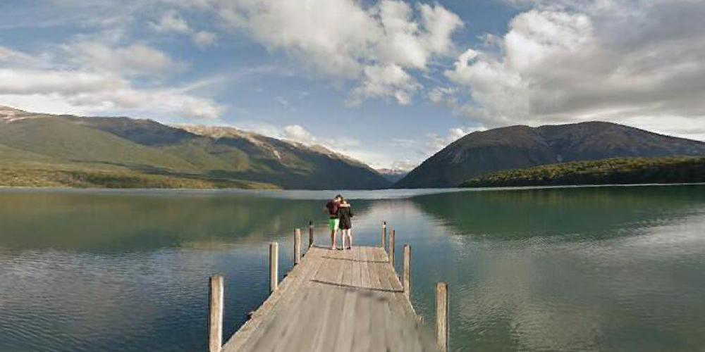 dock with couple embracing at the end of it, a large lake around them and mountains and clouds in the distance.