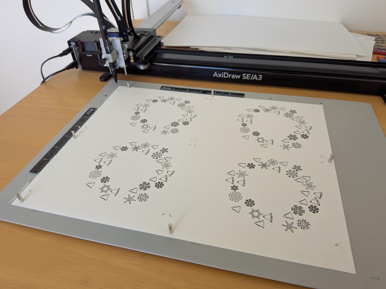axidraw pen plotter drawing four wreath tree cards and with the words "happy holidays" or "merry christmas" across the top of each